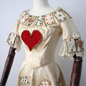 May include: Cream-colored dress with playing card accents and a large red heart on the bodice. The dress features playing card details around the neckline, sleeves, and skirt, with gold trim and lace accents. The dress is displayed on a mannequin.