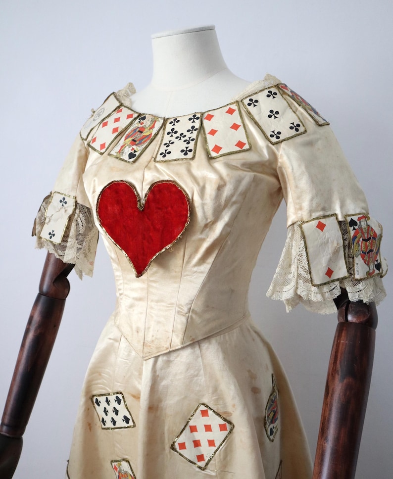 May include: Cream-colored dress with playing card accents and a large red heart on the bodice. The dress features playing card details around the neckline, sleeves, and skirt, with gold trim and lace accents. The dress is displayed on a mannequin.
