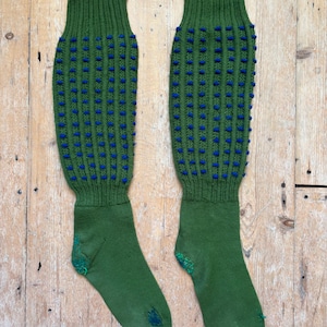Könnte beinhalten: Ein Paar grüne Strickstrümpfe mit blauen Bommeln und Bündchen. Die Socken haben ein strukturiertes Muster und werden auf einer Holzoberfläche präsentiert. Die Socken sind kniehoch.