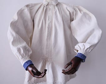 May include: Cream-colored vintage blouse with a round, eyelet-trimmed collar and pleated front. The long sleeves have puffed shoulders and blue patterned cuffs. The blouse has a lace trim at the hem.