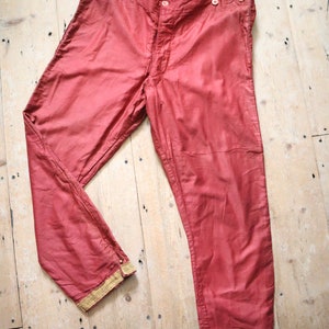 Op de afbeelding: Antieke rode broek met hoge taille en knoopsluiting. De broek heeft een licht glanzende uitstraling en is afgezet met goudkleurig kant aan de manchetten. Het kledingstuk wordt gepresenteerd op een licht houten oppervlak.