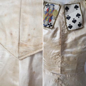 May include: Cream-colored vintage garment with playing card accents. The bodice features a Jack of Clubs and a Seven of Spades card, trimmed with gold. The fabric shows age spots, adding to its antique aesthetic. The garment has a button-down front.
