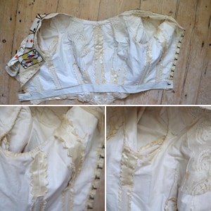 May include: An antique white corset with intricate detailing. The corset features decorative lace, satin ribbon accents, and a playing card patch on the sleeve. The garment is displayed on a wooden surface.