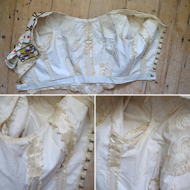 May include: An antique white corset with intricate detailing. The corset features decorative lace, satin ribbon accents, and a playing card patch on the sleeve. The garment is displayed on a wooden surface.