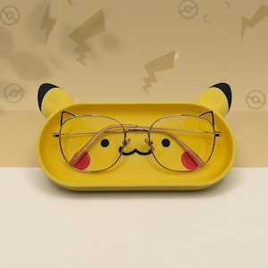 Könnte beinhalten: Ein gelbes Brillenablagefach im Pikachu-Design mit schwarzen Ohren und einem Cartoon-Gesicht. Das Fach enthält eine Brille mit goldfarbenem Rahmen und roten Akzenten. Der Hintergrund ist hellbeige mit Pikachu-Themen-Blitz- und Pokéball-Designs.