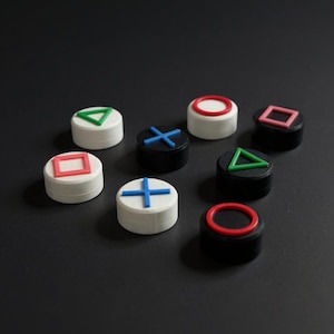 Könnte beinhalten: Eine Sammlung farbenfroher, knopfförmiger Objekte, die an Videospiel-Controller-Tasten erinnern. Die Knöpfe weisen eine Vielzahl von Formen und Farben auf, darunter Rot, Grün, Blau und Weiß, vor einem dunklen Hintergrund.