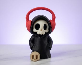 May include: A small figurine of a skeleton wearing a black hooded robe and bright red over-ear headphones. The figure has a white skull face and is standing behind a separate, smaller, light beige skull resting on the ground in front of it.