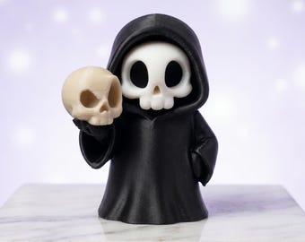 May include: A small figurine of a grim reaper character wearing a black hooded robe. The figure has a white skull-shaped head with large black eye sockets and holds a separate, smaller beige-colored skull in its right hand.
