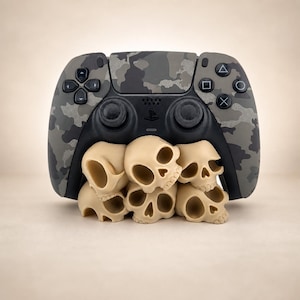 Könnte beinhalten: Ein Videospiel-Controller mit grau-schwarzem Tarnmuster, der auf einer Ansammlung von fünf kleinen, beigen, stilisierten Totenkopf-Figuren ruht. Der Controller verfügt über Standard-Steuerungstasten, zwei Analogsticks und vier Aktionstasten.
