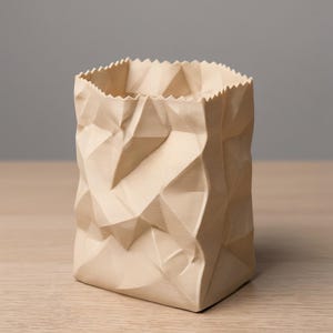 Puede incluir: Un jarrón de cerámica beige con diseño de bolsa de papel arrugada. Tiene base cuadrada y borde superior dentado. La superficie texturizada simula papel plegado. Se encuentra sobre una superficie de madera clara.