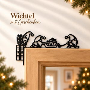 Pode incluir: Uma decoração de canto de porta em metal preto com três gnomos com presentes e um trenó. O texto Wichtel mit Geschenken está impresso acima do design. O ornamento está posicionado no canto superior de uma moldura de porta de madeira.