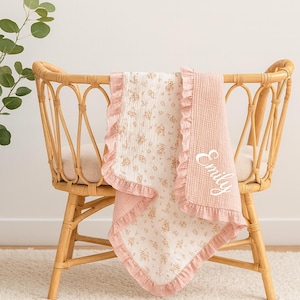 May include: A pink and white baby blanket with a floral pattern and ruffled edges. The blanket is draped over a wicker bassinet. The name "Emily" is embroidered on the blanket in white script.