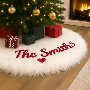 May include: A white, fluffy Christmas tree skirt with the embroidered text "The Smiths" in red, along with a red heart. The skirt is under a decorated Christmas tree with gold ornaments and string lights. Several wrapped gifts are placed around the tree.