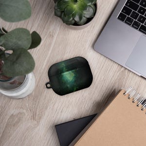 Op de afbeelding: Een zwarte en groene Airpods-hoes met een galaxy-thema en een kleine lus om een ​​sleutelhanger aan te bevestigen.