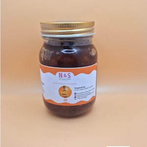 May include: A jar of Sunny Delight Pineapple jam with a gold lid and a white label with orange accents. The label includes the text "N&S" and "JAM". The jar is filled with a dark, reddish-brown preserve.