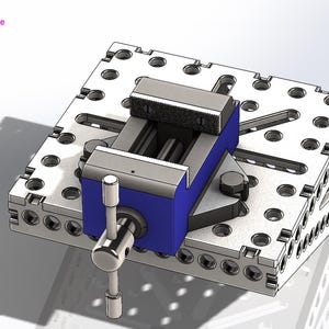 May include: A silver and blue metal vise mounted on a perforated metal base. The vise has a blue body and a silver handle. The base is square with numerous holes. The image has the text "Ariana France" in the upper left corner.