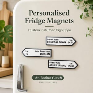 May include: Three rectangular fridge magnets designed like Irish road signs with white backgrounds, black borders, and black text. The magnets display the names of Irish towns in both Irish and English, along with distance markers in kilometres.