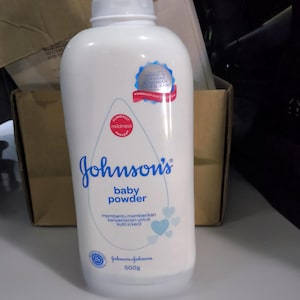 May include: A white plastic bottle of Johnson's baby powder with a blue cap. The label features the text Johnson's baby powder, 500g, and includes blue heart graphics, a red circular seal, and a blue circular seal with Indonesian text.