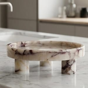 May include: A round, marble tray with three cylindrical legs. The tray is a light beige color with dark purple veining. The tray is sitting on a white marble surface.