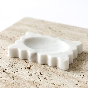 May include: A white marble soap dish featuring a rectangular shape with scalloped, wavy edges and a smooth, recessed center basin. The stone displays subtle grey veining throughout its polished surface, providing a clean and functional design.