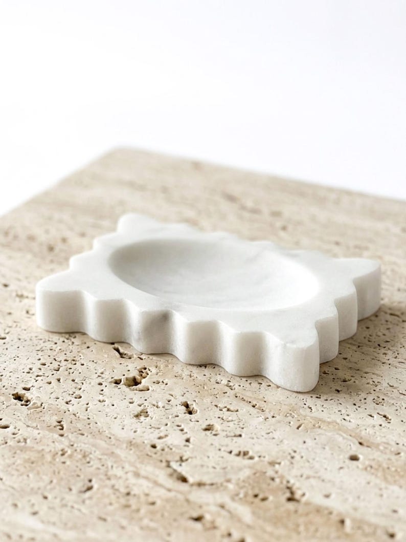 May include: A white marble soap dish featuring a rectangular shape with scalloped, wavy edges and a smooth, recessed center basin. The stone displays subtle grey veining throughout its polished surface, providing a clean and functional design.