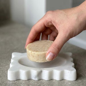 May include: A round, beige, textured soap bar held above a square, white marble soap dish with a scalloped, wavy edge. The soap bar has visible flecks of botanical ingredients throughout its surface, and the marble dish features a smooth, polished finish with subtle grey veining.