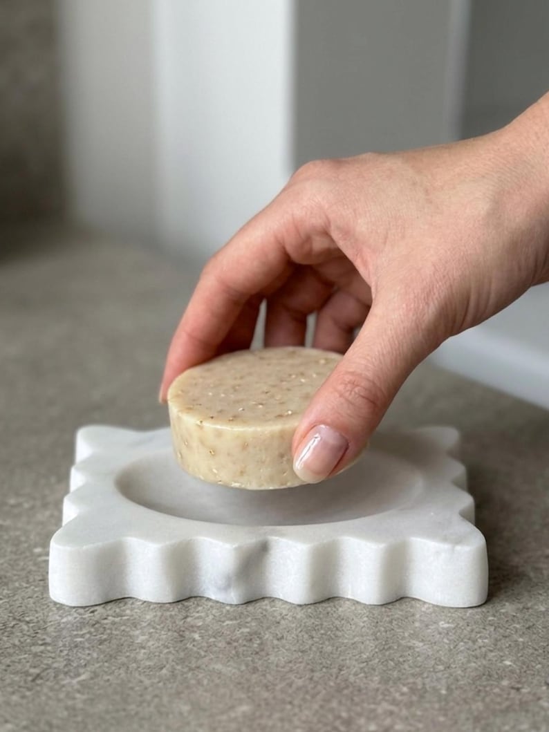 May include: A round, beige, textured soap bar held above a square, white marble soap dish with a scalloped, wavy edge. The soap bar has visible flecks of botanical ingredients throughout its surface, and the marble dish features a smooth, polished finish with subtle grey veining.