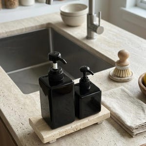 May include: Two glossy black square soap dispensers with black pump mechanisms, one larger than the other, resting on a rectangular beige travertine stone tray. The dispensers have a smooth, reflective finish and are positioned side by side.