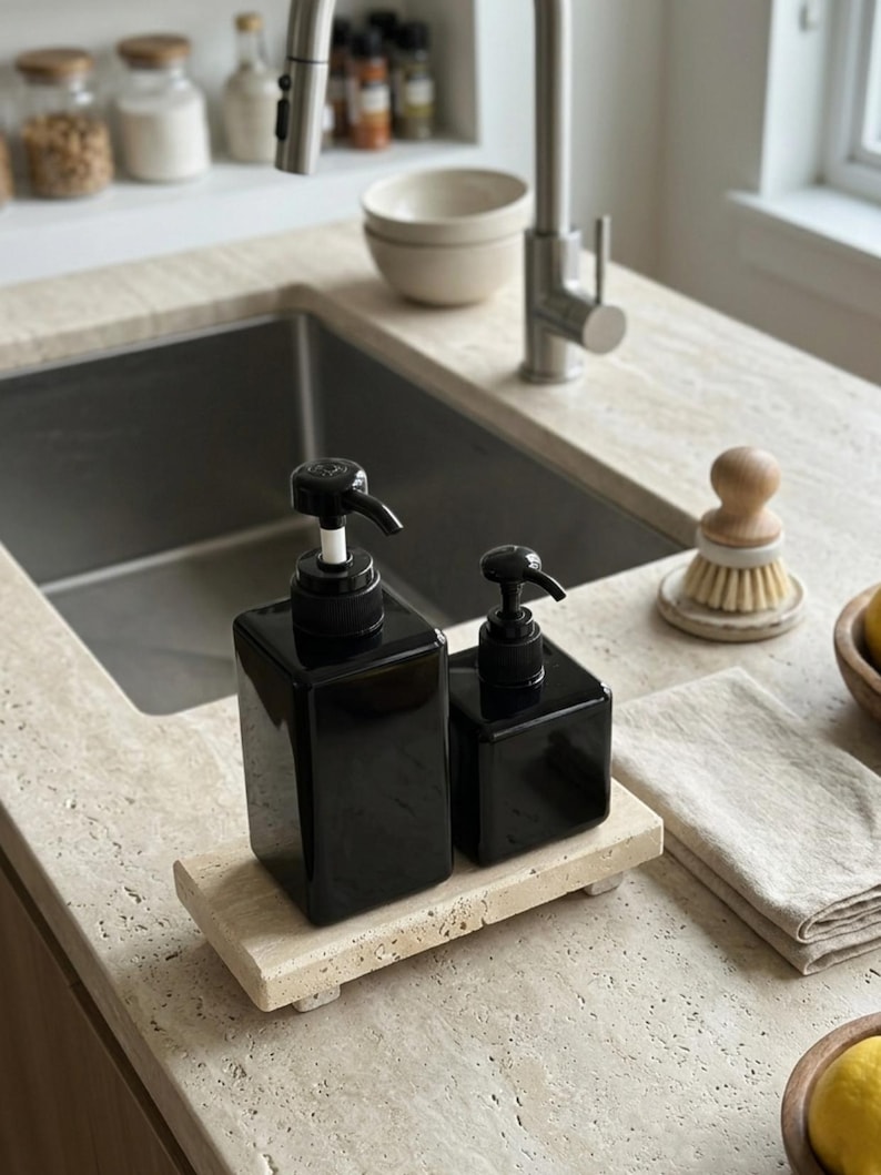May include: Two glossy black square soap dispensers with black pump mechanisms, one larger than the other, resting on a rectangular beige travertine stone tray. The dispensers have a smooth, reflective finish and are positioned side by side.