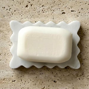 May include: A rectangular white bar of soap resting on a white marble soap dish. The soap dish features a scalloped, wavy edge design. The bar of soap is smooth, solid, and opaque, centered perfectly on the decorative marble tray surface.