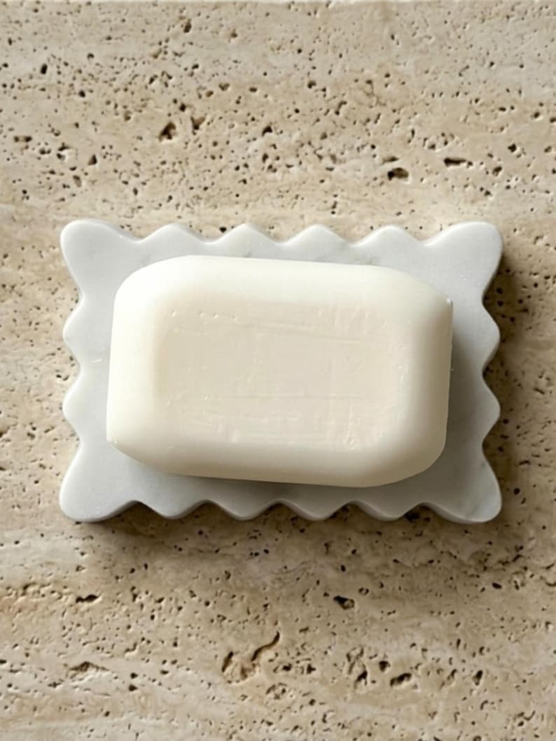 May include: A rectangular white bar of soap resting on a white marble soap dish. The soap dish features a scalloped, wavy edge design. The bar of soap is smooth, solid, and opaque, centered perfectly on the decorative marble tray surface.