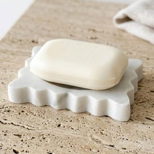 May include: A rectangular off-white bar of soap resting on a white marble soap dish with a scalloped edge. The soap dish has a smooth, polished surface and a distinct wavy perimeter, providing a stable base for the solid, smooth-textured soap bar.