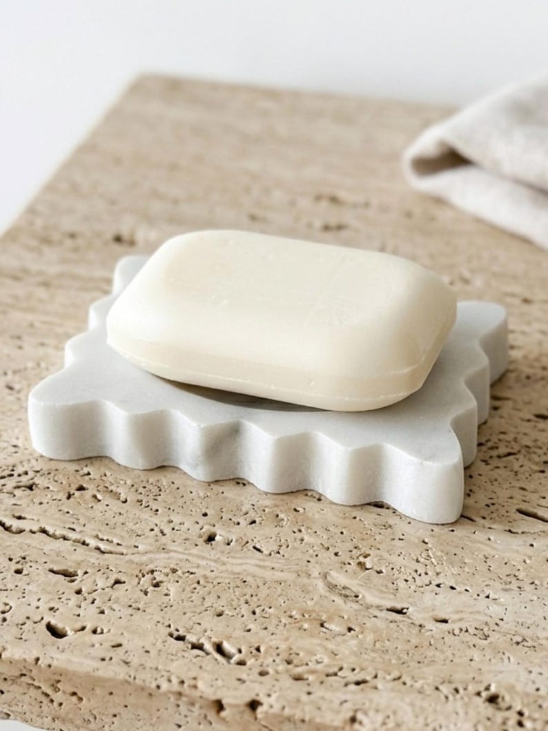 May include: A rectangular off-white bar of soap resting on a white marble soap dish with a scalloped edge. The soap dish has a smooth, polished surface and a distinct wavy perimeter, providing a stable base for the solid, smooth-textured soap bar.