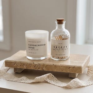 May include: A white wax candle in a glass jar labeled Sandalwood Rose and a clear glass bottle filled with wooden safety matches, both resting on a rectangular beige travertine stone riser with four small feet and a decorative lace ribbon.
