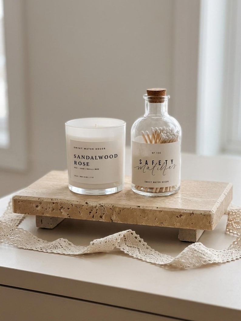 May include: A white wax candle in a glass jar labeled Sandalwood Rose and a clear glass bottle filled with wooden safety matches, both resting on a rectangular beige travertine stone riser with four small feet and a decorative lace ribbon.