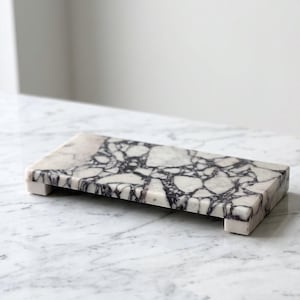 May include: A rectangular marble riser featuring a white base with prominent, dark purple and black veining in a brecciated pattern. The piece is elevated on four small, square marble feet, showcasing a polished, smooth surface texture.