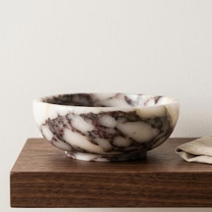 May include: A small, round marble bowl with a white base and a swirling pattern of burgundy and gray. The bowl sits on a dark brown wooden shelf, with a neutral-colored cloth partially visible on the right.