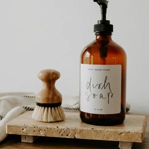 May include: An amber glass soap dispenser with a black pump and a white label reading "SWEET WATER DECOR dish soap 16 FL OZ" next to a round wooden dish brush with light-colored bristles, both resting on a rectangular beige stone tray.