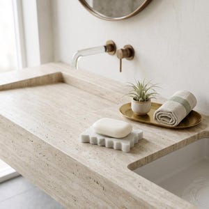 May include: A beige travertine stone bathroom sink featuring a wall-mounted faucet with a clear spout and brass handle. A white bar of soap rests on a scalloped white tray, while a small potted succulent and a rolled beige towel sit on a gold tray.