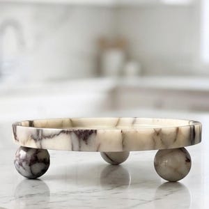 May include: A circular marble tray featuring a smooth, flat surface with cream and dark purple veining. The tray is elevated by three spherical marble feet that match the stone pattern of the main circular base, creating a raised display platform.