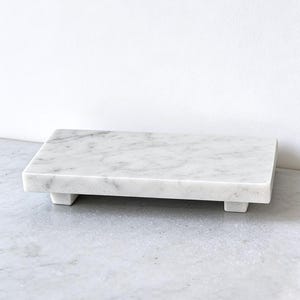 May include: A rectangular white marble tray featuring natural grey veining throughout the stone surface. The tray is elevated by four small, short block feet at each corner, providing a stable base for this polished, solid stone display piece.