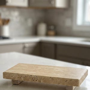 May include: A rectangular beige travertine stone riser with a porous, textured surface and natural veining. The platform is elevated by four small, matching stone feet, providing a stable base for display purposes on a flat surface.