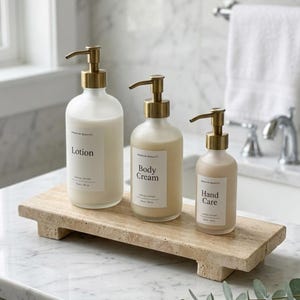 May include: Three frosted glass pump bottles with gold-toned dispensers, labeled Lotion, Body Cream, and Hand Care, arranged on a rectangular beige travertine stone tray. Each bottle features a white label with black text and a pump mechanism.