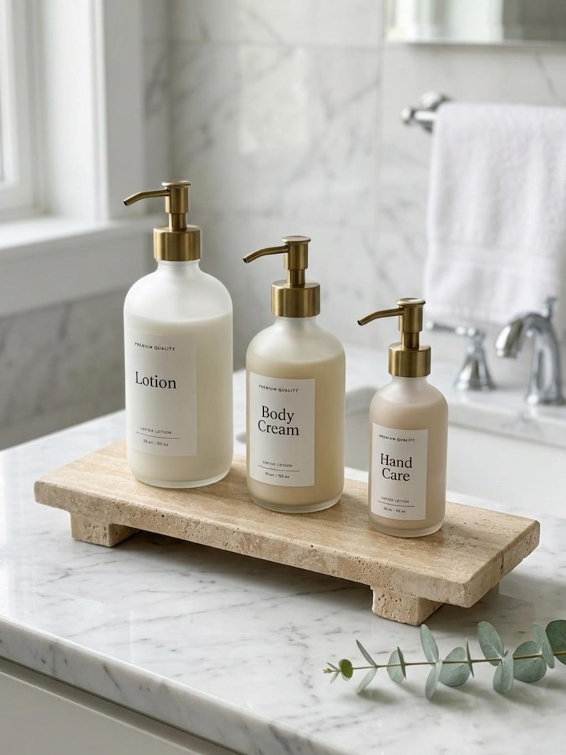 May include: Three frosted glass pump bottles with gold-toned dispensers, labeled Lotion, Body Cream, and Hand Care, arranged on a rectangular beige travertine stone tray. Each bottle features a white label with black text and a pump mechanism.