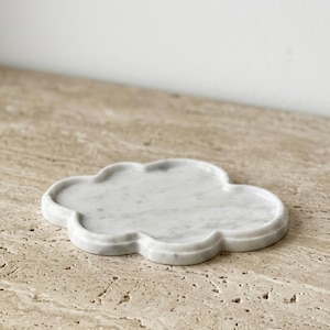 May include: A white marble tray shaped like a cloud with a smooth, polished surface and subtle grey veining throughout. The tray features a raised, rounded edge that follows the scalloped outline of the cloud shape, creating a shallow basin.