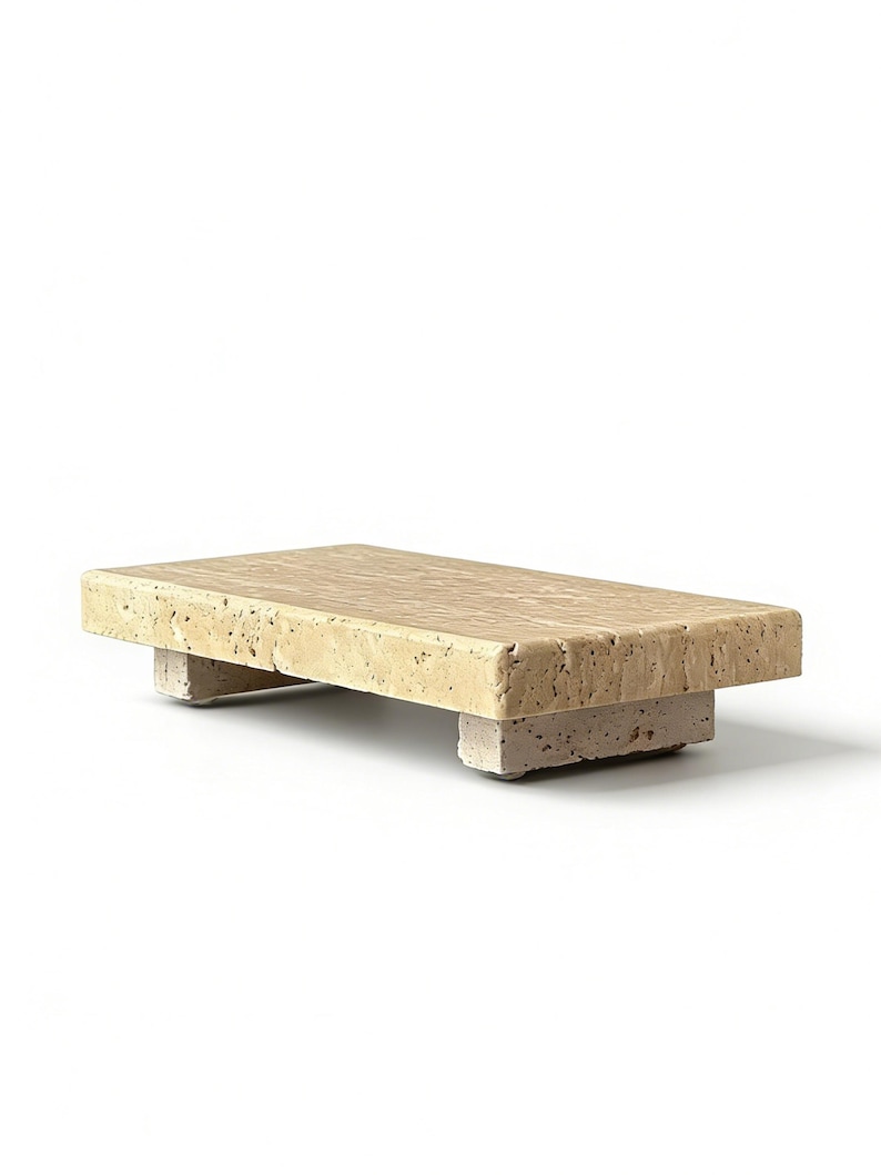 May include: A rectangular beige travertine stone riser with a porous, textured surface. The platform is supported by two thick, matching stone block feet, creating a low-profile stand with a natural, earthy appearance and visible stone grain.