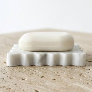 May include: A smooth, solid white bar of soap resting on a square, white marble soap dish. The soap dish features a scalloped, wavy edge design with subtle grey veining throughout the stone material, providing a clean and functional bathroom accessory.