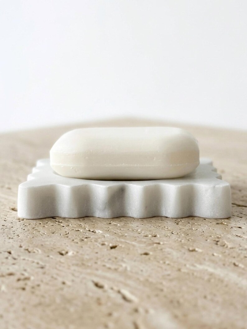 May include: A smooth, solid white bar of soap resting on a square, white marble soap dish. The soap dish features a scalloped, wavy edge design with subtle grey veining throughout the stone material, providing a clean and functional bathroom accessory.