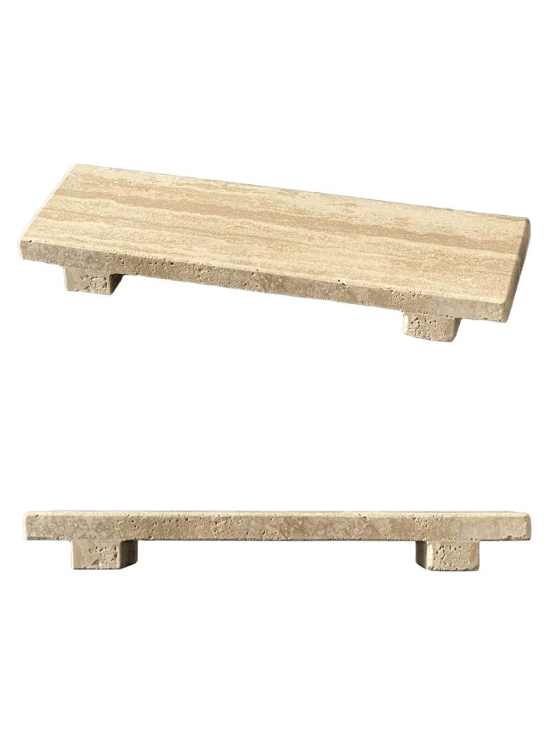 May include: A rectangular beige travertine stone riser with a porous, natural texture. The platform features a flat top surface supported by two small, block-shaped feet at each end, providing a slightly elevated display base for various objects.