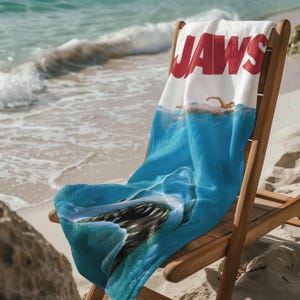 May include: A beach towel featuring the movie title "JAWS" in red letters on a white background. The towel depicts a swimmer in the ocean with a shark beneath the surface, in shades of blue.